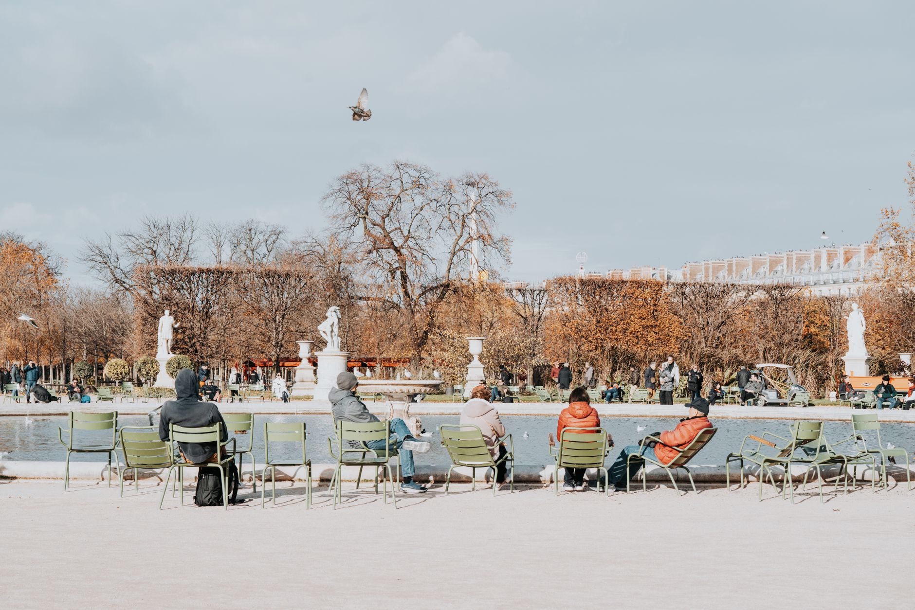 People sitting on green chairs around a round pond in the Tuileries under an overcast sky, with autumn trees and classical statues behind