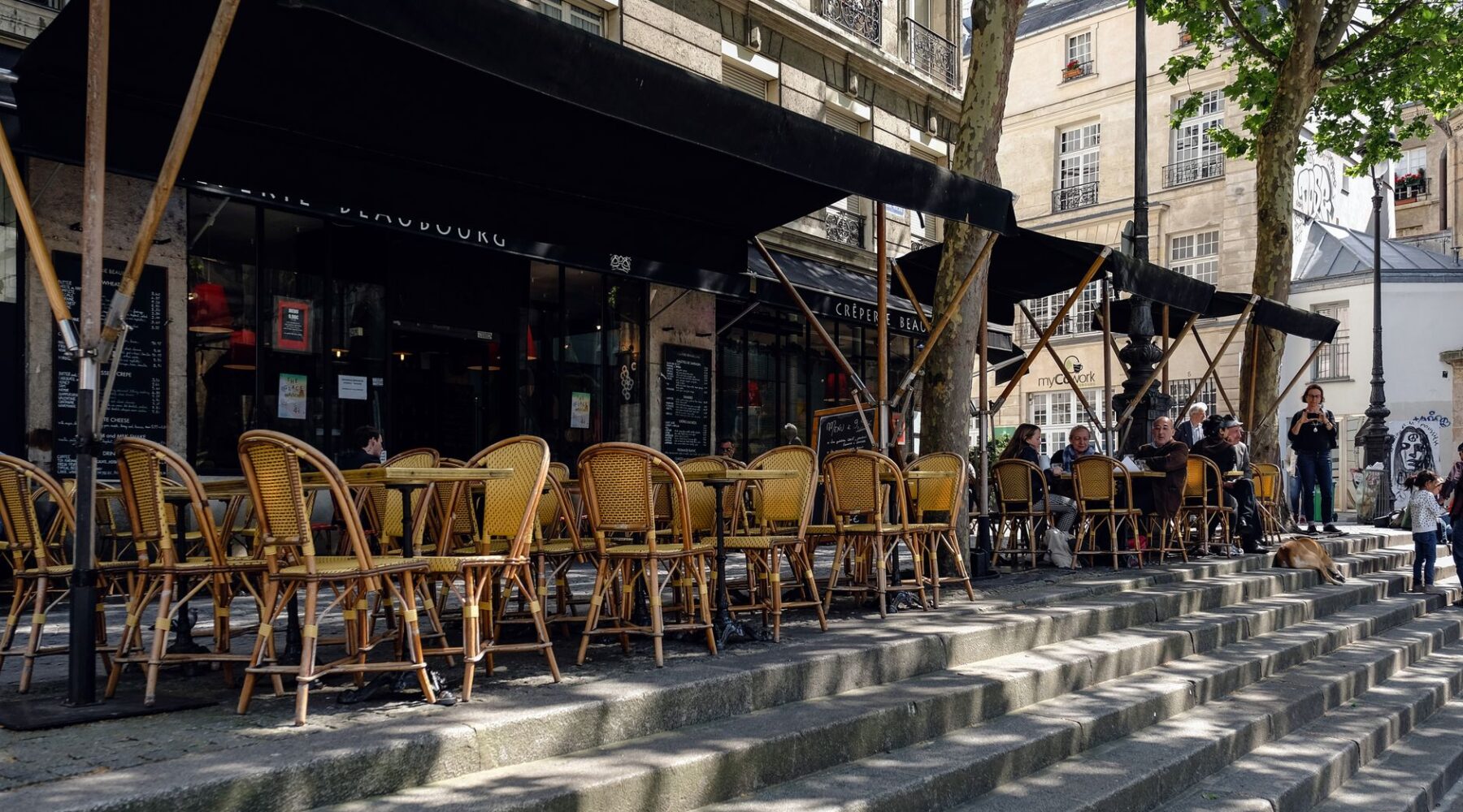 Rattan cafe chairs and tables under black awnings on a sunny Paris pavement terrace