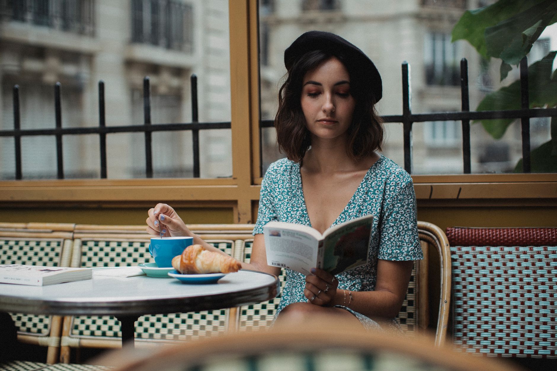 A woman sitting alone at a Parisian café terrace with a coffee and a croissant, reading a paperback on a rattan chair