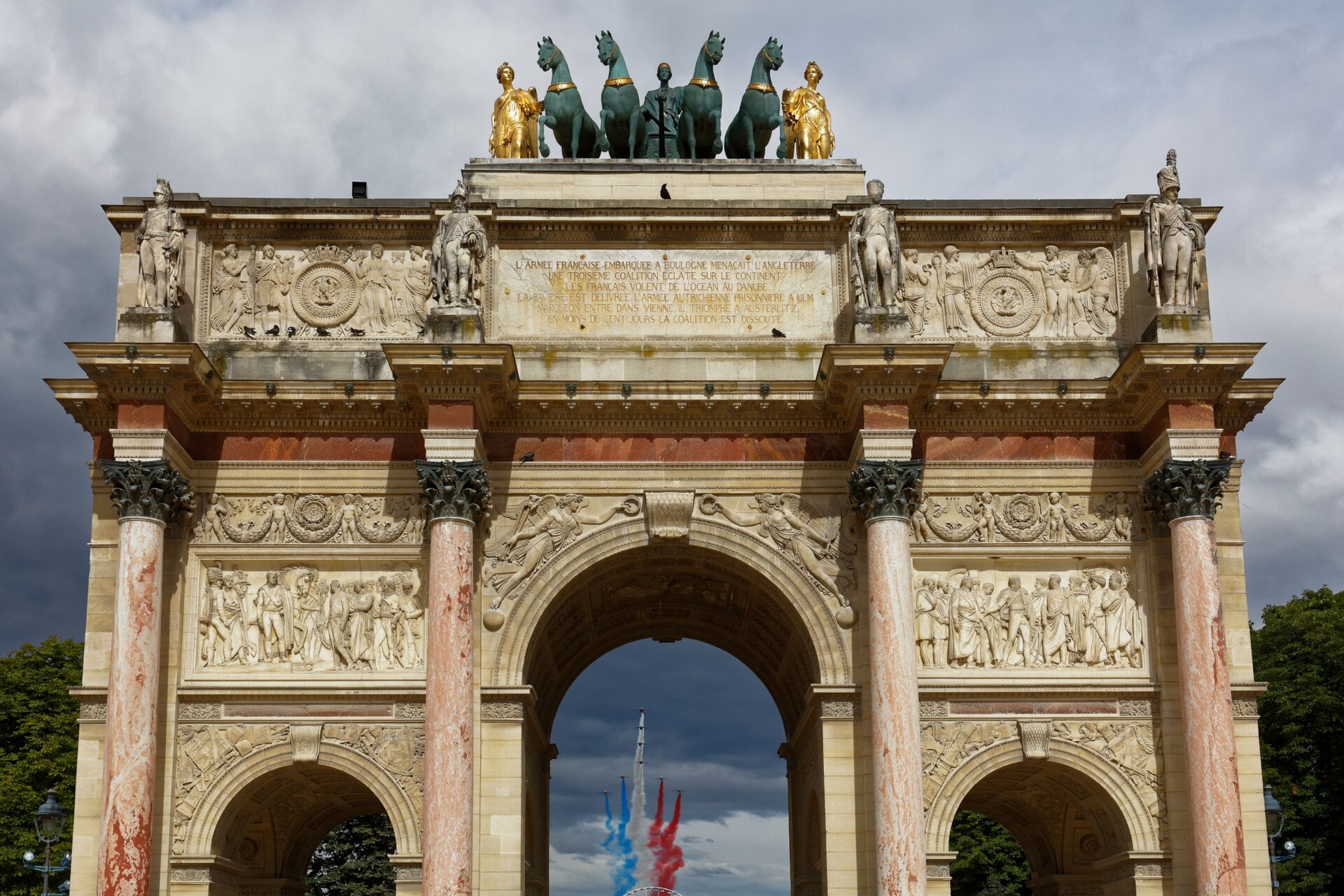 The Arc de Triomphe du Carrousel under a cloudy sky, with trails of blue, white and red smoke from the Bastille Day flypast visible through its central arch
