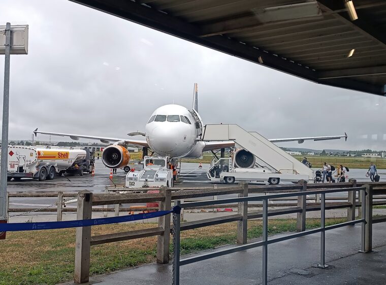 Passengers walking across the wet tarmac to board a white Airbus A319 by its stairs at Beauvais–Tillé airport, seen from behind the terminal railings
