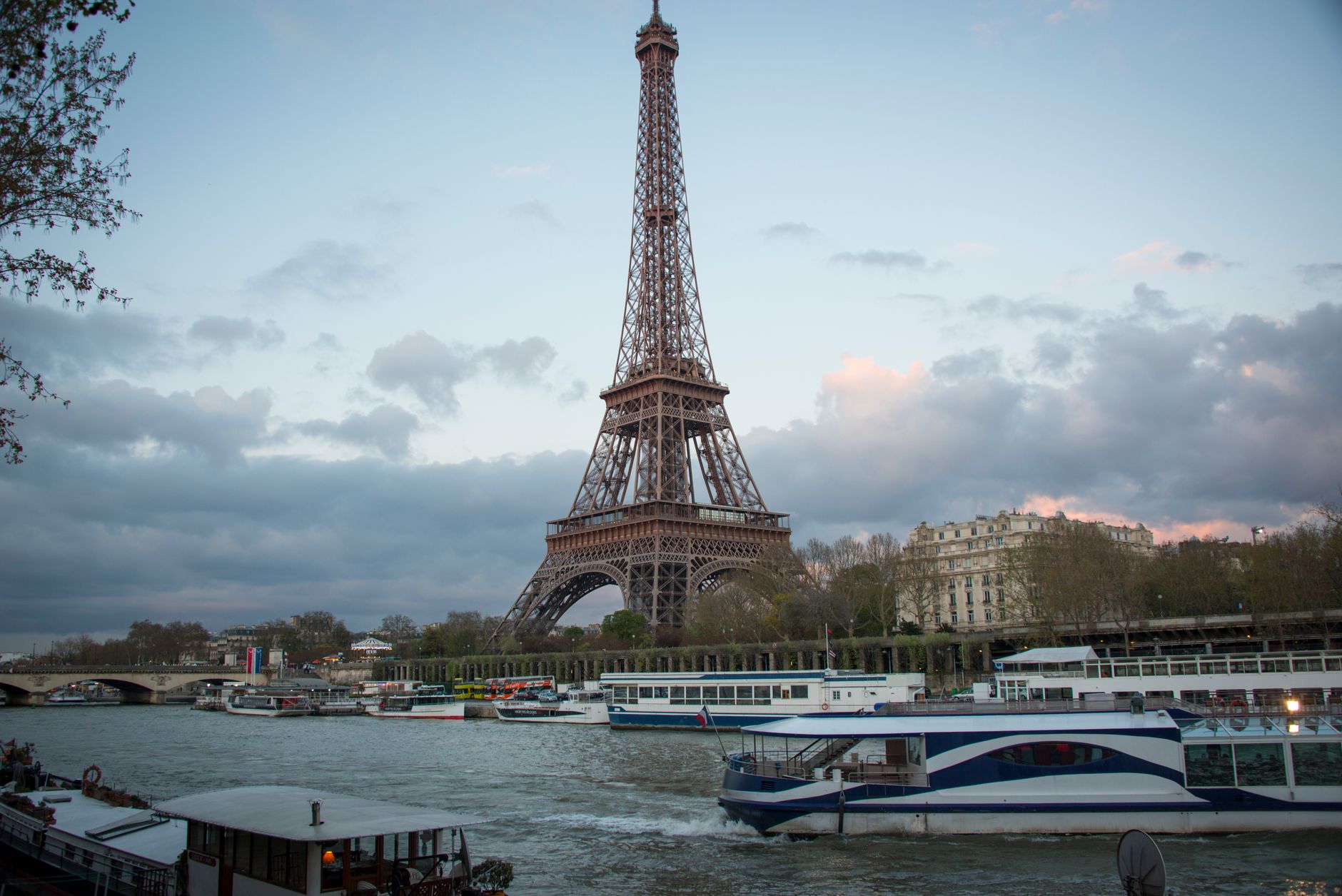 The Eiffel Tower at dusk above the Seine, with sightseeing boats moored along the river and pink clouds in the sky
