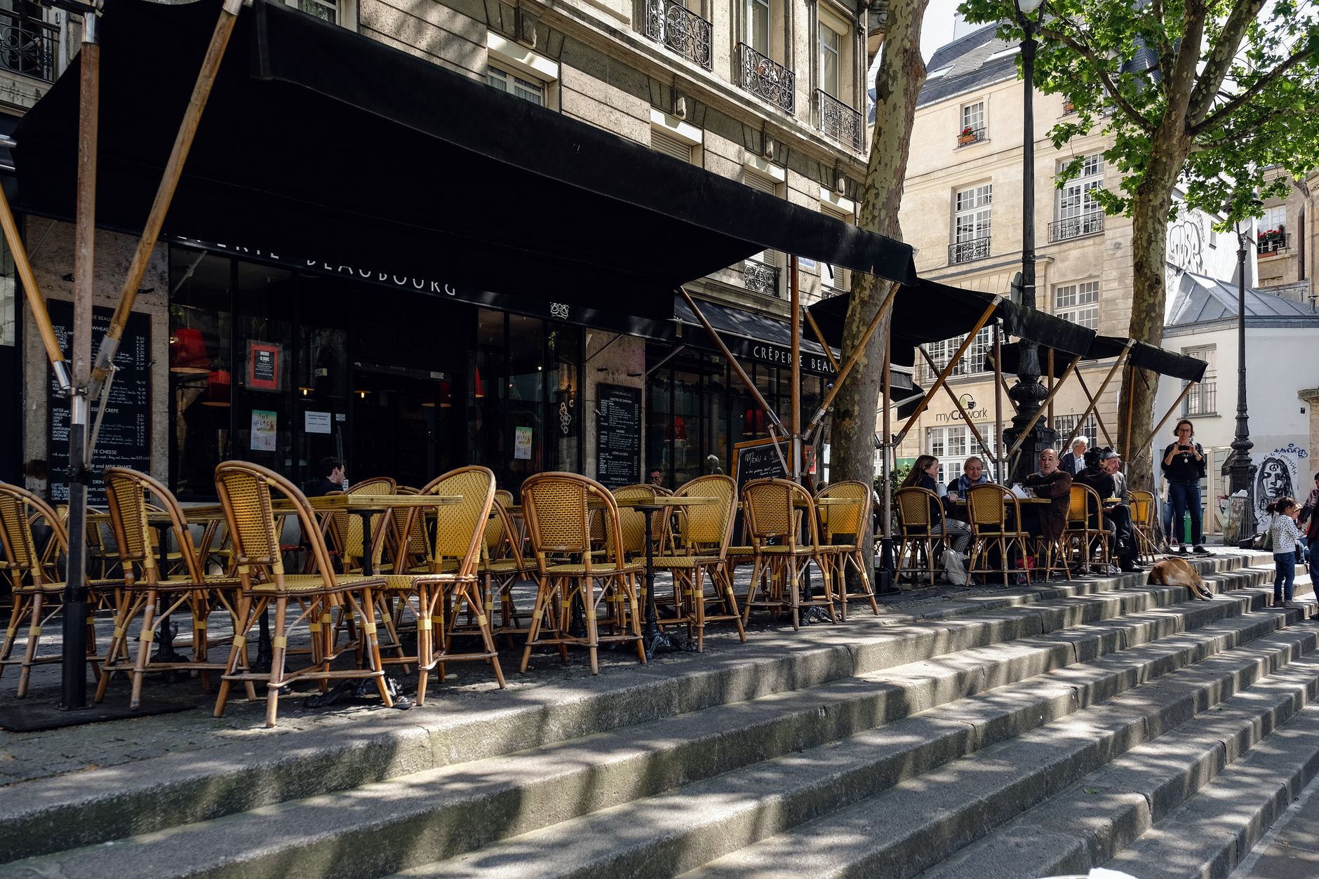 Rows of rattan café chairs on a stepped terrace under black awnings on a sunny day in Paris, with a few people sitting at the far end