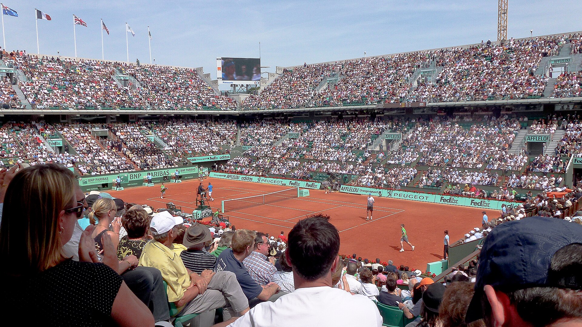 Spectators in the stands watching a match on the red clay of Court Philippe-Chatrier at Roland-Garros under a blue sky