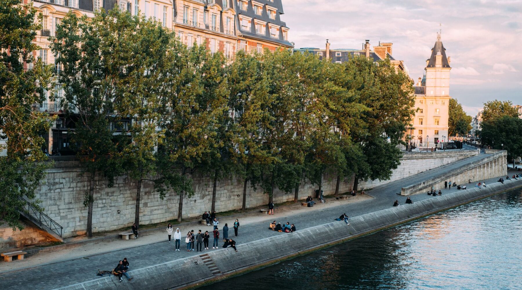 People sitting along the lower quay of the Seine beneath plane trees and tall Parisian buildings in warm evening light