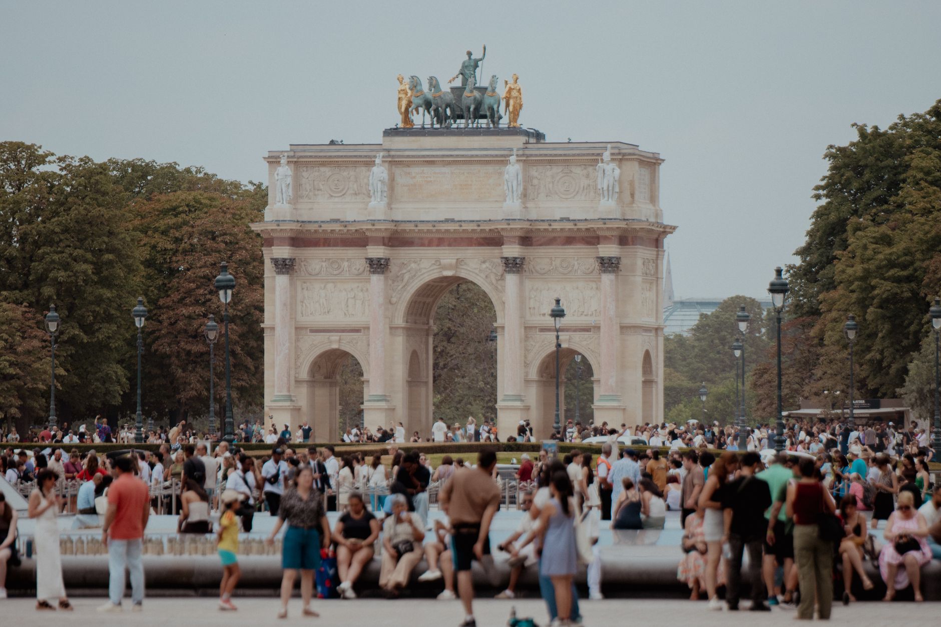 Summer crowds sitting and standing around a pond in the Jardin des Tuileries, with the Arc de Triomphe du Carrousel and trees behind